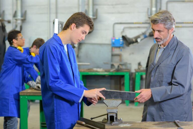 Apprenti en blouse bleue et son formateur travaillant une pièce métallique à l'étau en atelier